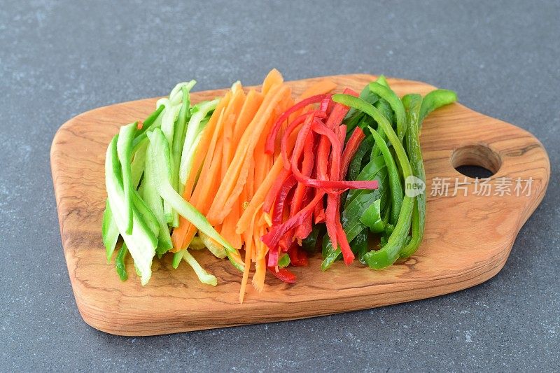 鲜黄瓜、胡萝卜、红绿甜辣椒在灰色抽象背景的橄榄木砧板上切成条状。分步烹饪