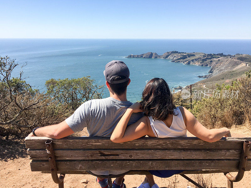 一对年轻夫妇坐在长椅上欣赏美妙的海景