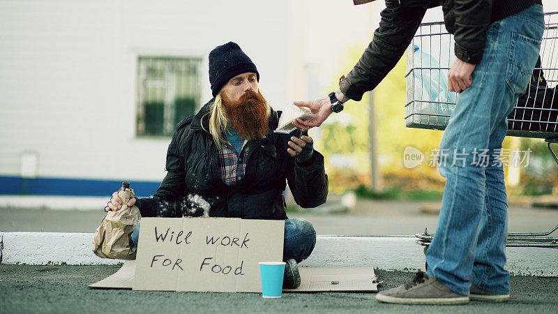 年轻人帮助无家可归的人，给他一些食物，而乞丐喝酒，坐在附近的购物车在街上