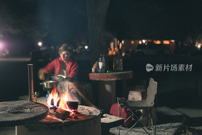 妇女晚上用柴火和烤肉设备做饭。前景是帐篷和椅子。在非洲国家公园探险。健美的形象。