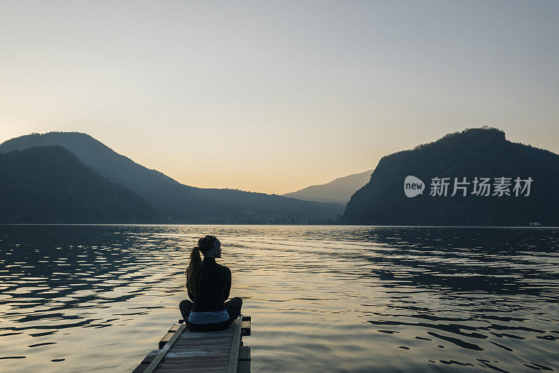 一个女人坐在湖边的码头边，看着日出越过山脉和森林