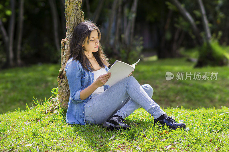 年轻快乐的女人坐在草地上，靠在一棵树上，欣赏着一本书