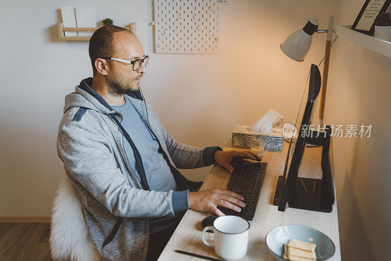 一个白人在他的家庭办公室里用电脑工作一边喝着他的早上咖啡