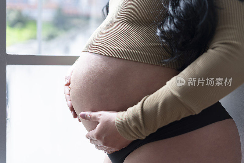 一个妈妈的肚子在窗口的特写，爱抚她的肚子，因为她等待着她的新婴儿在她温暖的土地睡衣的到来。