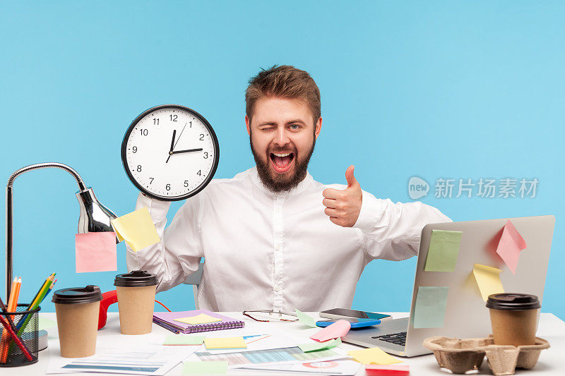 快乐微笑的男人办公室职员，胡子在白衬衫显示拇指和眨着眼睛拿着挂钟，按时完成工作，时间管理