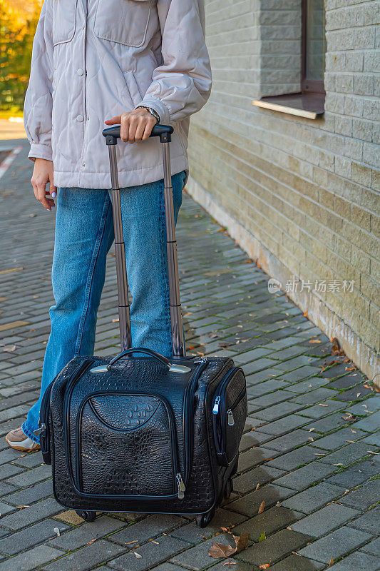在阳光明媚的天气里，一名身着休闲服装、提着手提箱的年轻女子在火车站的机场以一堵砖墙为背景摆出姿势