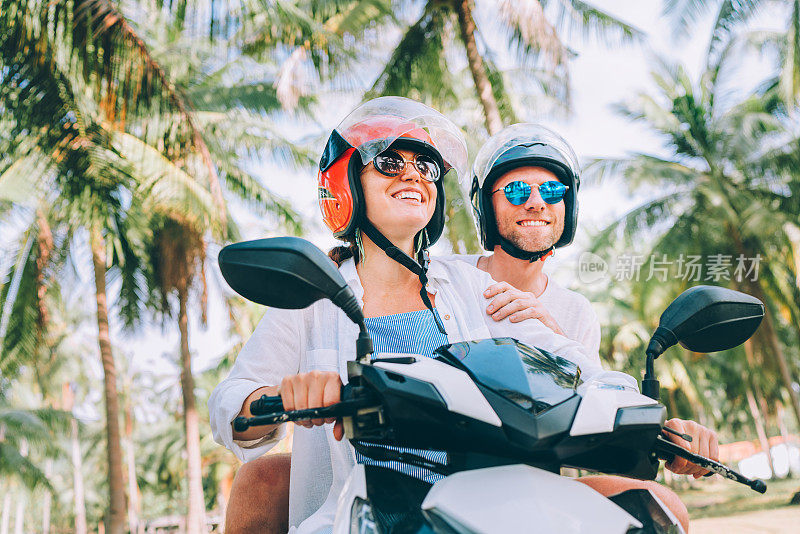 在泰国苏梅岛的热带度假中，戴着安全头盔骑着摩托车的一对快乐的旅行者在棕榈树下微笑