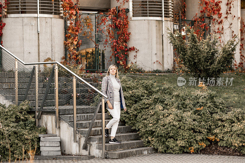 秋日，一名迷人的中年妇女穿着格子夹克和灰色卷领毛衣走下混凝土楼梯。