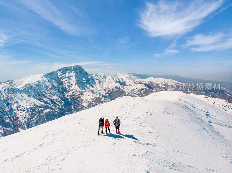 图为，在冬季登上高海拔雪山顶峰的成功登山队手持登山杆，看着摄像机拍摄的无人机照片