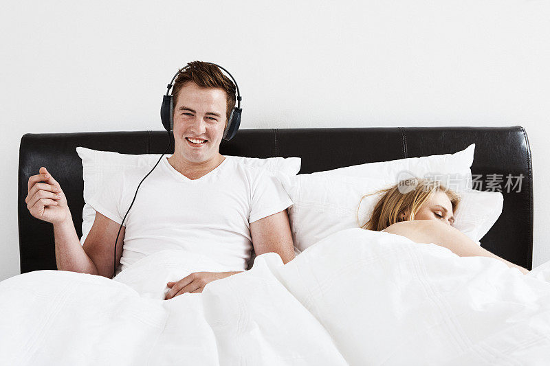 当妻子或女友睡在他旁边时，男人坐在床上戴着耳机听音乐，脸上露出微笑
