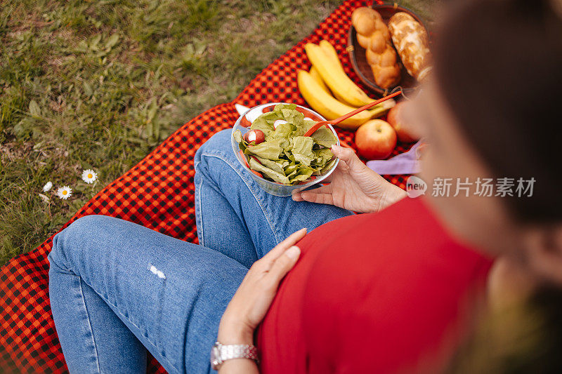 年轻漂亮的孕妇坐在红色的毯子上，吃着混合蔬菜沙拉。