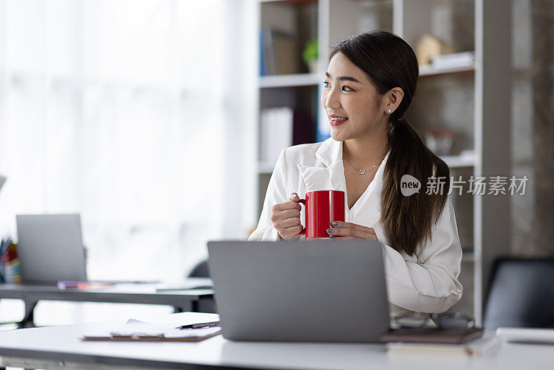 一位亚洲女性的经理在她的办公桌上容光焕发地喝着咖啡。年轻的亚洲女性坐在桌子前，用她的笔记本电脑上网，