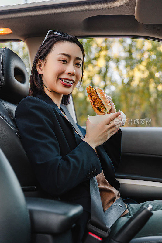 亚洲女人在车里休息，吃汉堡。一个年轻成功快乐的女人正在开车。