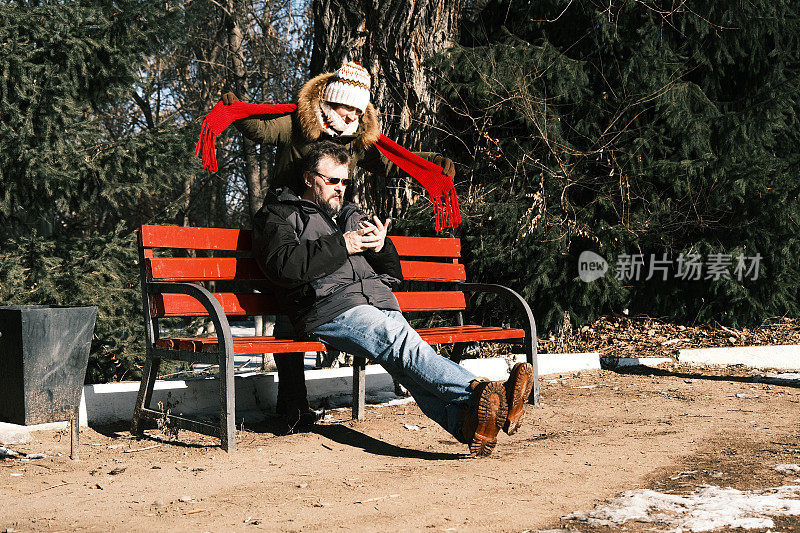 女人在冬季公园里玩，而她的丈夫坐在那里玩手机