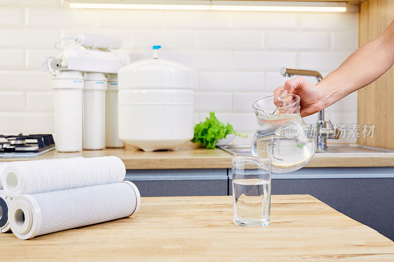 在厨房里，一名妇女正在用手把纯净水从罐子里倒进玻璃杯里。反渗透过滤器的背景。保健和美容。Hydratation。