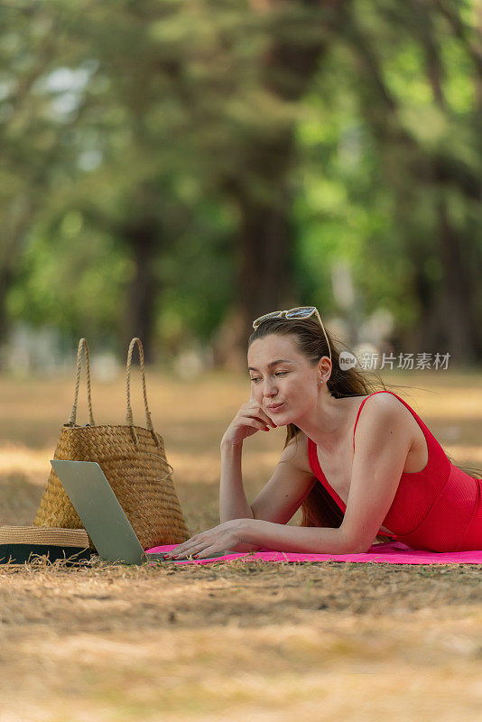 美丽的白人女性性感的女人在红色泳装比基尼戴着太阳镜躺在地上工作在她的电脑笔记本电脑放松在树林里的海滩