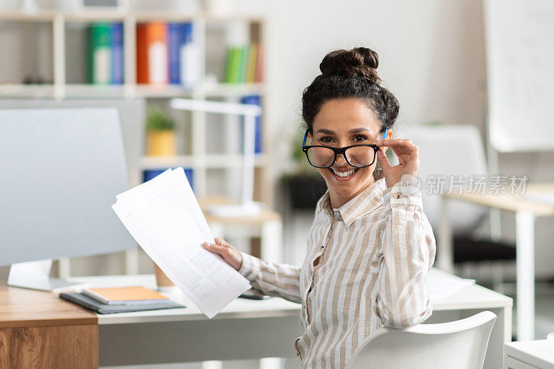 女企业家在办公室办公桌前处理文件的肖像，摘下眼镜，对着镜头微笑