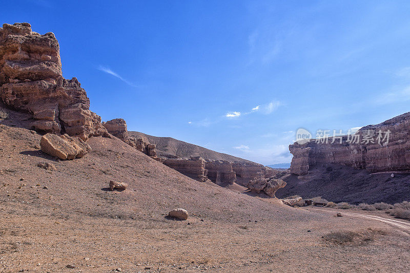 哈萨克斯坦沙漠中巨大的查林峡谷。岩石的视野与相邻的道路。炎炎夏日，枯黄的草和烈日映衬着蓝天和罕见的云。由风形成的地质层