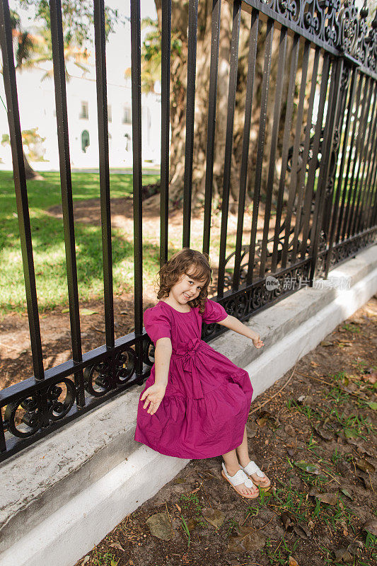 可爱的古巴裔美国三岁小女孩，棕色卷发，棕色眼睛和橄榄色肤色，穿着亮紫色连衣裙，蝴蝶结和白色凉鞋，站在佛罗里达州棕榈滩湖小径的黑色栏杆前