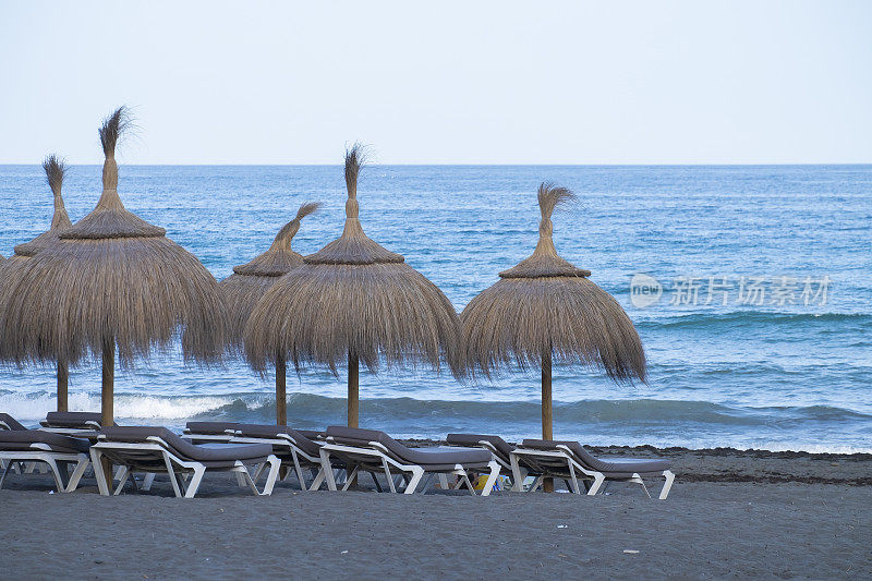 地中海岸边的稻草沙滩伞和舒适的日光浴躺椅。