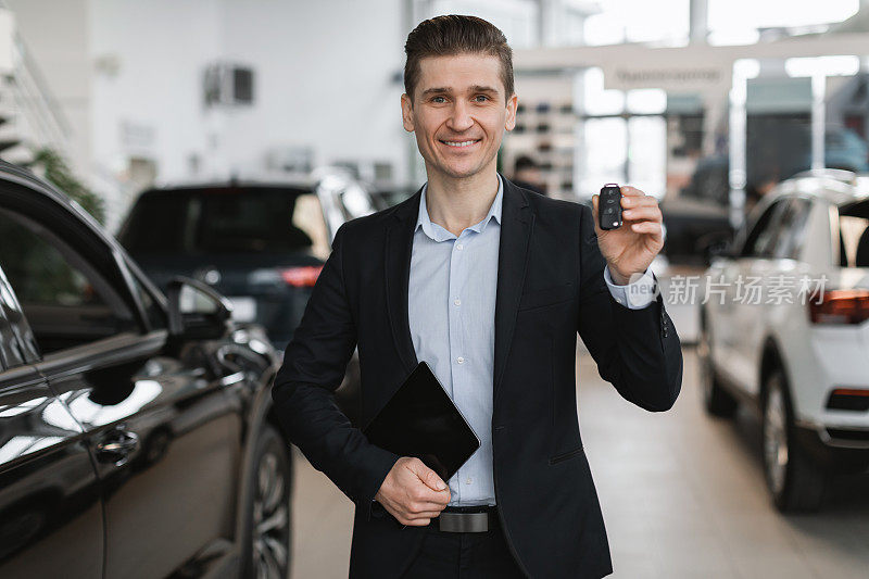 年轻英俊的汽车销售员在相机展示汽车钥匙，在经销商销售汽车