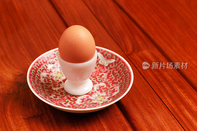 一个鸡蛋在一个白色钢化玻璃蛋杯上瓷碟放置在红木餐桌上。没有人