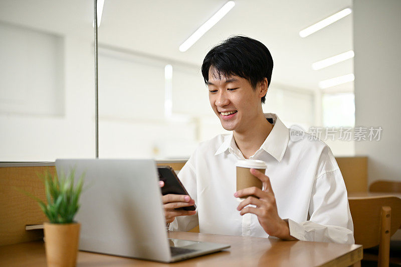 一个快乐的亚洲男人坐在咖啡店里，一边喝着咖啡，一边玩着智能手机。