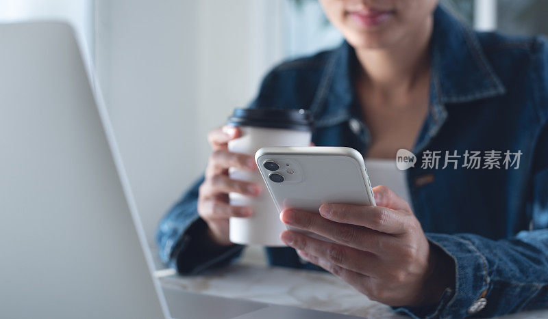 年轻聪明的亚洲女商人坐在桌子旁，喝着咖啡，用手机，桌上放着笔记本电脑，在家庭办公室工作时休息一下