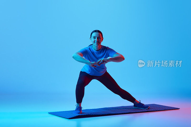 积极，微笑的成熟女人站在健身matt上，在霓虹灯的蓝色工作室背景下训练