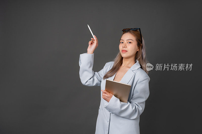 留长发的亚洲女性高管或公司员工戴时尚眼镜，穿蓝色西装。在你身边放一个平板电脑，在灰色背景的工作室里拍一张照片