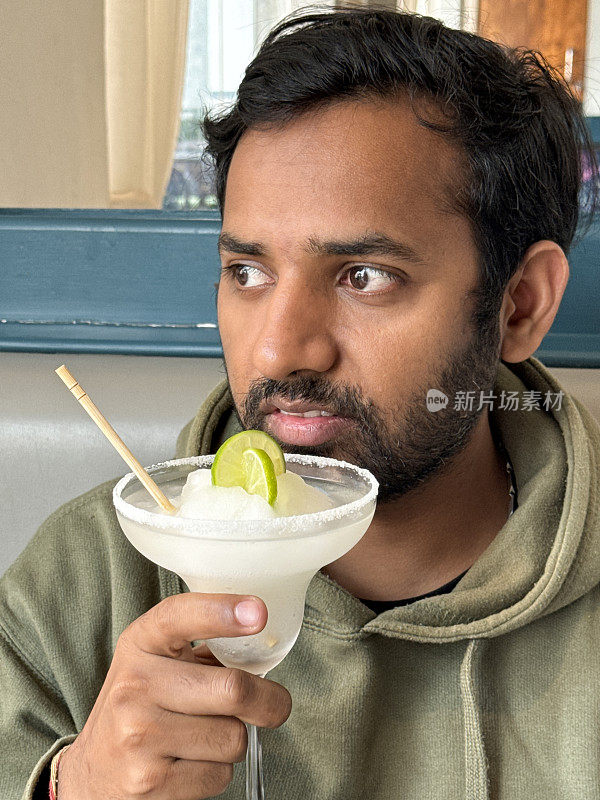 餐厅场景的特写图片，印度人从冷冻玛格丽塔龙舌兰鸡尾酒，柠檬片装饰，木制搅拌棒，重点在前景的盐边喝玛格丽塔杯