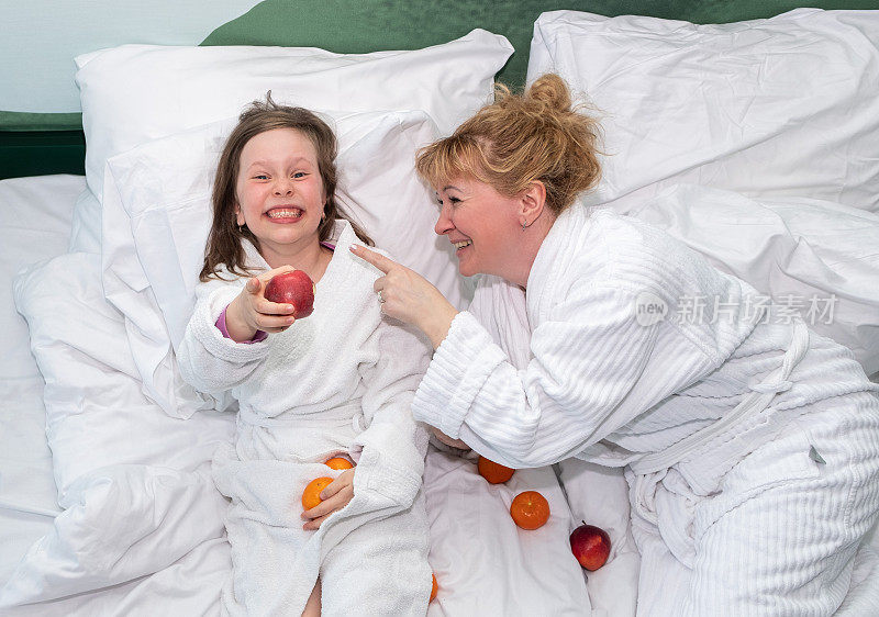 一个年轻的女人和一个7岁的小女孩躺在床上开心地笑着。一个女人和一个孩子吃水果。