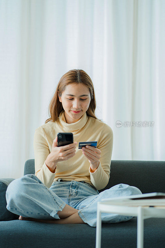 开朗的亚洲女性使用智能手机，网上购物，手持信用卡，支付，坐在家里的沙发上网上银行和电子商务应用垂直图片