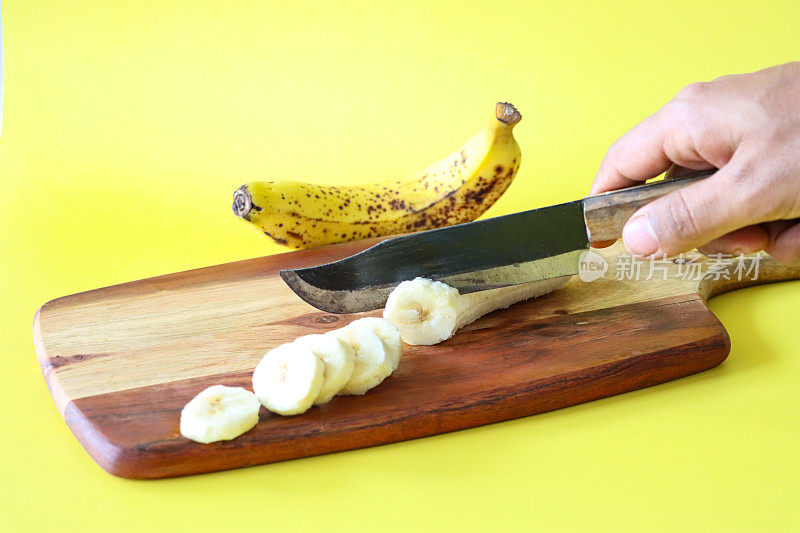 不可识别的人用菜刀在木制砧板上切香蕉的特写图像，整个未削皮的成熟水果和去皮的切片，黄色背景，重点放在前景，复制空间