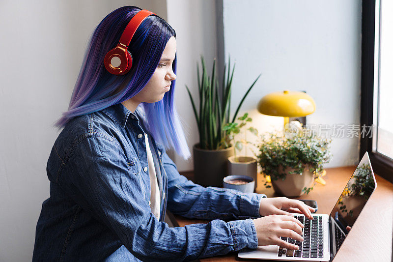 迷人的年轻拉丁裔女孩的肖像，蓝头发，蓝色牛仔衬衫，坐在咖啡馆里，在笔记本电脑上打字，还有复印空间。