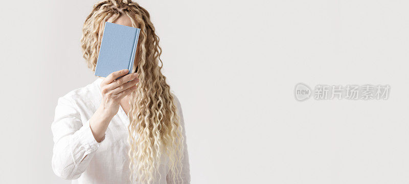 女子手持蓝色封面的纸质书在脸前，浅色背景，无脸潮流概念。年轻女性，长卷发，脏辫藏在抄写日记后面。教育的概念