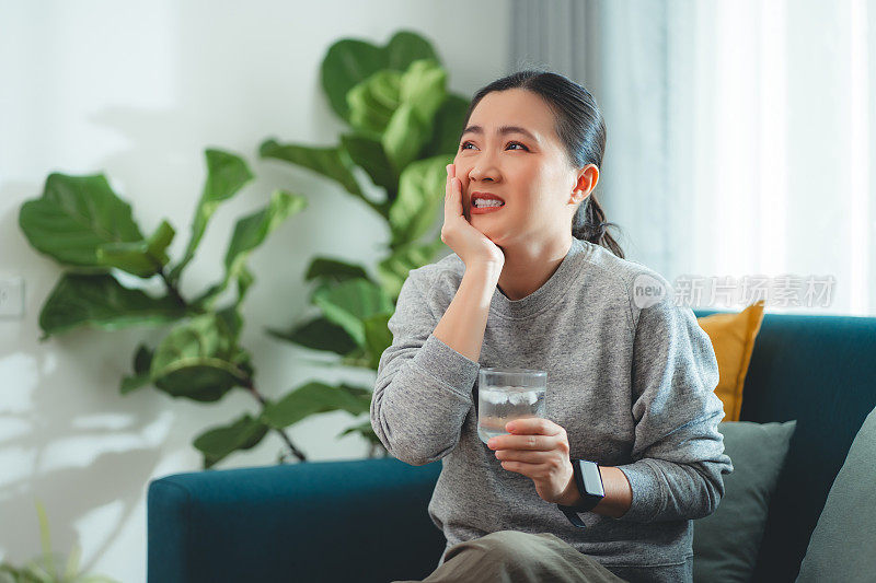 亚洲女性坐在客厅的沙发上，喝了凉水，摸了摸脸颊，就会感到牙齿敏感。