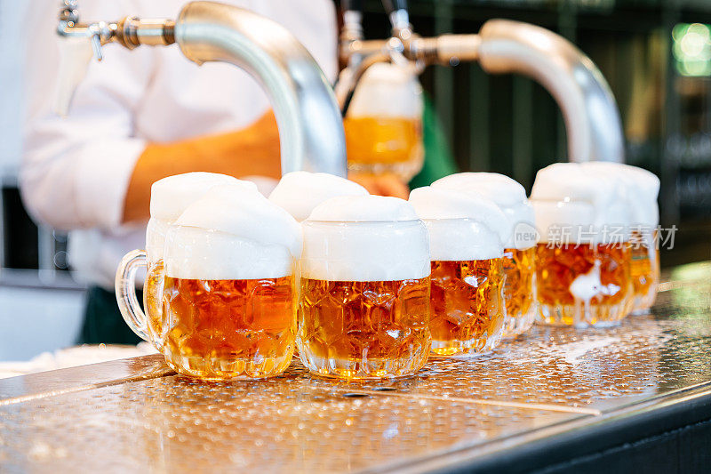 近距离拍摄的调酒师将冰镇啤酒倒入杯口起泡的玻璃杯，拍摄于一个带有金属台面的现代酒吧。