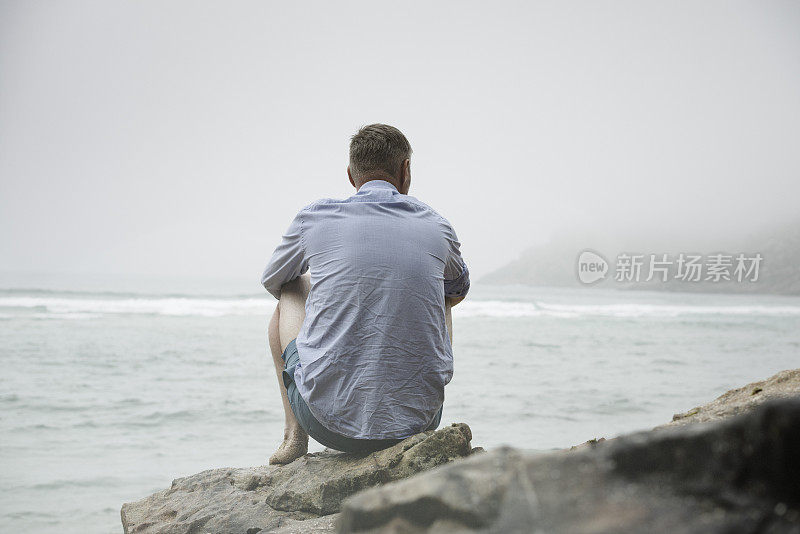 一个男人坐在岩石上，眺望着雾蒙蒙的海滩