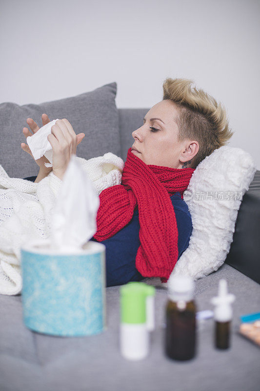 一个生病的年轻女子因感冒坐在家里的沙发上，她盖着一条毯子，擤着鼻子