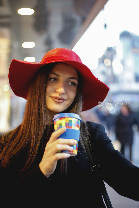 年轻女子戴着红帽子，在城市街道上使用智能手机和喝咖啡