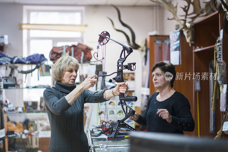 迷人的金发成熟，50岁的女人在小狩猎商店检查弓，与女售货员的协助。加里宁格勒，俄罗斯，东欧。