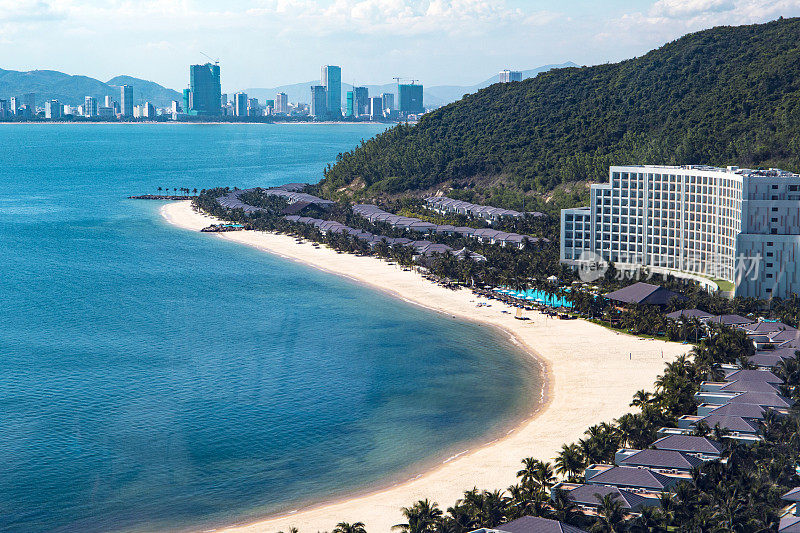 沙滩上有棕榈树，白色的伞，沙滩椅在蓝色的海边，以山和城市为背景