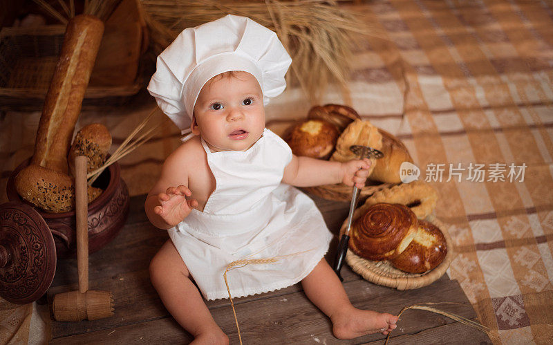 快乐的小男孩穿着厨师服坐在厨房的桌子上，旁边的篮子里装着美味的面包卷、小圆面包和面包。本空间