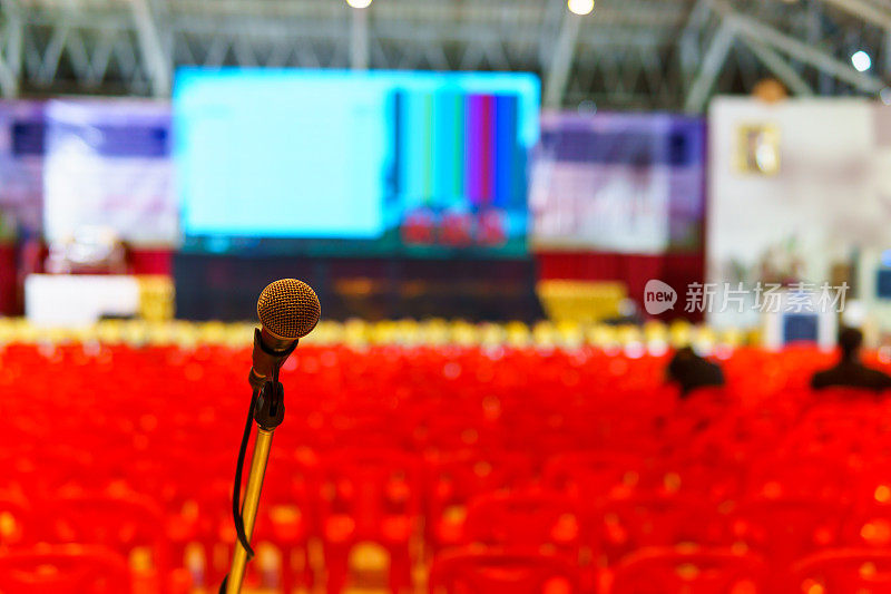 近距离的麦克风在研讨会大厅扬声器或歌手模糊排红色塑料椅子和大led显示屏背景。为文本复制空间。讲师、演讲者、研讨会概念。