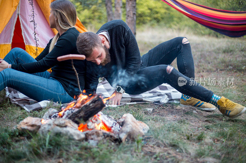 漂亮的一对，围坐在篝火旁。背景是一个帐篷，那人用一根棍子弹火。