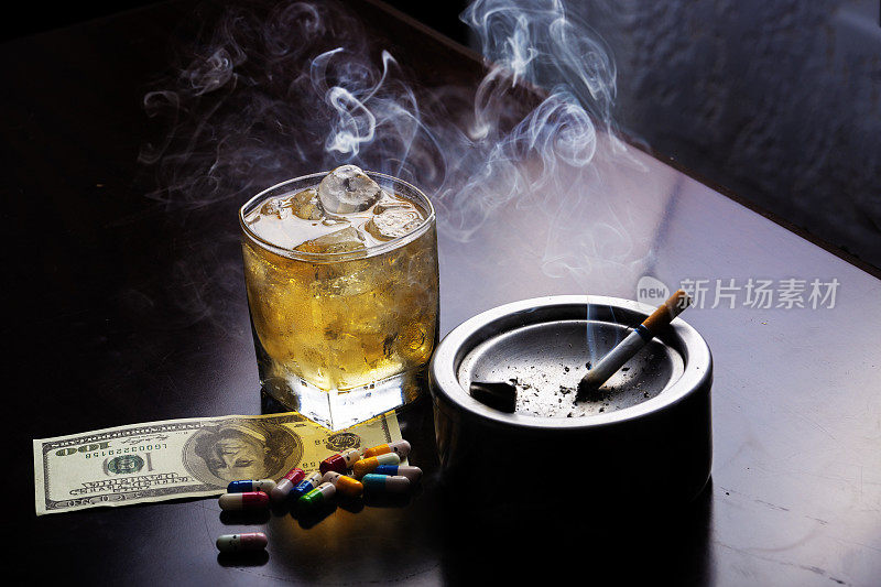 烟灰缸旁边放着一杯酒还有满是烟雾的酒吧里装着100美元钞票的药丸