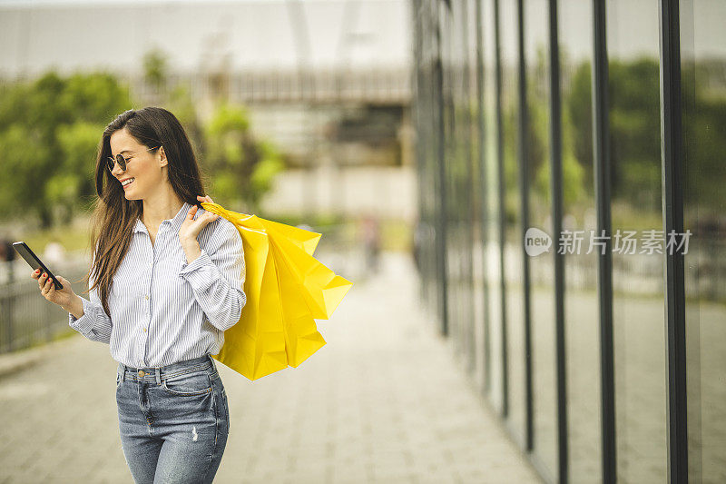 年轻漂亮的女人提着购物袋，用智能手机走路