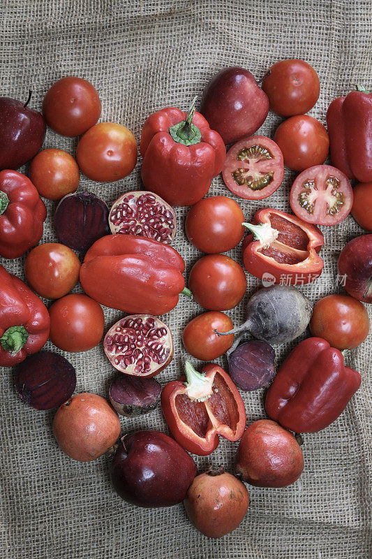 这是一堆心形的红色水果和蔬菜，包括甜椒、甜菜根、西红柿、苹果和石榴，放在灰色的粗棉布背景上，这是一种对心脏有益的饮食理念，是一种居高一下的观点