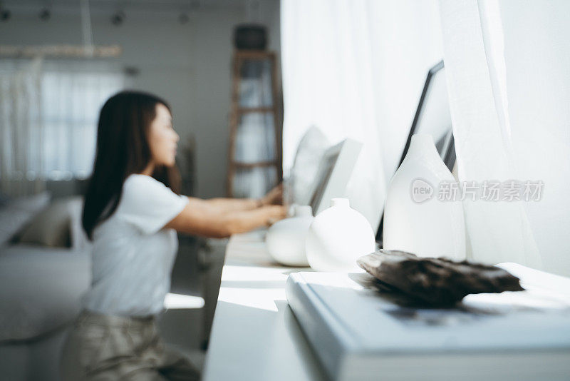 一个年轻的亚洲女人在家里窗户边的架子上装饰和整理相框和花瓶的散焦图像